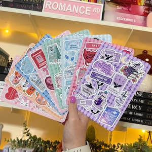 May include: A hand holds a stack of colorful, patterned stickers with various designs and text. The stickers feature themes like fantasy, books, and winter, with phrases such as "Book Dragon" and "Fantasy RD". The background includes a shelf with books.
