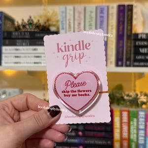 May include: A pink Kindle grip with the words "kindle grip" and "Please skip the flowers buy me books." The grip is heart-shaped and held in front of a bookshelf.