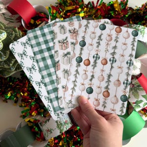May include: A hand holds a stack of Christmas-themed cards with various patterns. Designs include green bows, gingham, gift boxes, and ornaments. The cards are surrounded by festive ribbons and tinsel in red, green, and gold.