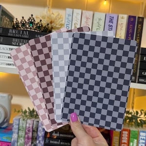 May include: A hand holds a fanned stack of four rectangular cards with checkered patterns in shades of pink, brown, gray, and navy blue. The cards are in front of a bookshelf with books and decorative items.