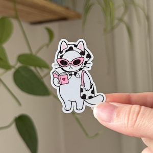 May include: A die-cut sticker featuring a white cat wearing a black and white cow-print headscarf and pink-rimmed sunglasses. The cat holds a small pink book with a heart icon and carries a matching cow-print shoulder bag on its side.