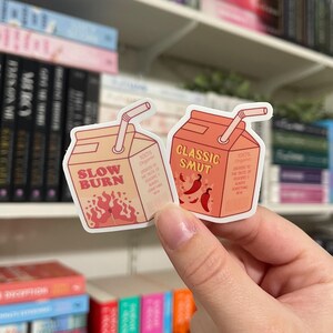 May include: Two cartoon juice box stickers with red and orange accents. The first sticker says "Slow Burn" with flames and the second sticker says "Classic Smut" with red chili peppers.