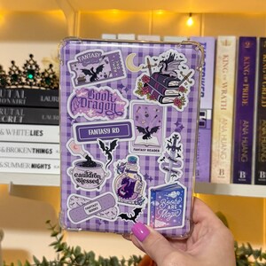 May include: A clear protective case with a purple gingham pattern and fantasy-themed stickers. Stickers include dragons, books, potion bottles, and the text "Book Dragon", "Fantasy RD", and "Books are Magic".