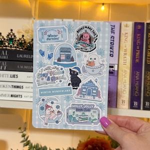 May include: A sticker sheet with a light blue checkered background and winter-themed illustrations. Designs include a snow globe, a cat in a Santa hat, and the text "Winter Carnival." The sheet is held by a person with pink nail polish.