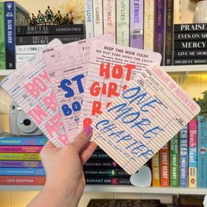 May include: A collection of vintage-style library card prints with bold text. The cards feature phrases like "Boy in love", "Hot Girl Reads", and "One More Chapter" in pink, blue, and red. The background includes books and a camera.