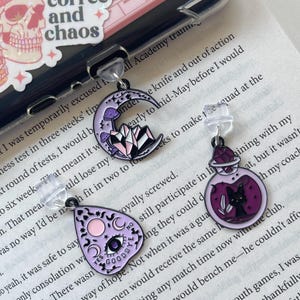 May include: Three purple and black enamel pins with different designs. The first pin is a crescent moon with a crystal and mushroom. The second pin is a purple teardrop with a crescent moon, an eye, and the words "Yes" and "No" with "Goodbye" underneath. The third pin is a purple potion bottle with a black cat and leaves.