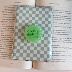 May include: E-reader case with a green and white chequered design. A green circular pop socket with the text "do not disturb I'm reading" is attached. The e-reader is open on a book.