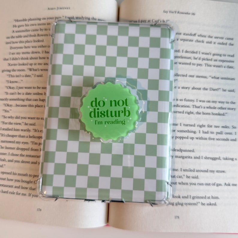 May include: E-reader case with a green and white chequered design. A green circular pop socket with the text "do not disturb I'm reading" is attached. The e-reader is open on a book.
