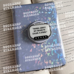 Könnte beinhalten: Eine rechteckige Handyhülle mit hellvioletter holografischer Oberfläche und einem runden Zubehörteil. Das Zubehör ist weiß mit schwarzem Rand und trägt den Text: BOOK REVIEW, MORAL GRAY? MY FAVORITE COLOR. 5 STARS.