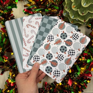 May include: A hand holds four rectangular cards with festive patterns. Designs include candy canes, checkered patterns, and gingerbread men on ornaments. The cards have a white background with gray and red accents, perfect for holiday decor.
