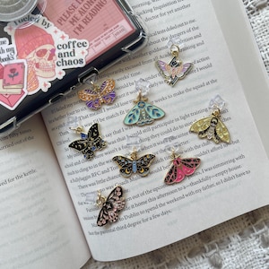 May include: Nine gold-toned butterfly bookmark charms with colorful enamel details. The bookmarks feature different butterfly designs with black, white, pink, blue, green, and yellow accents. The bookmarks are attached to clear plastic clips.