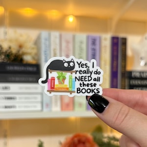 May include: A sticker featuring a black cat perched on a bookshelf with colorful books. The sticker reads "Yes, I really do NEED all these BOOKS." The sticker is held by a person with a black manicure, with a blurred background of more books.