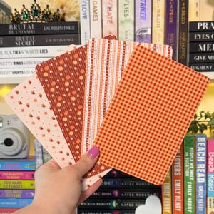 May include: A hand holds a collection of decorative cards with fall-themed patterns. The cards feature designs like acorns, stripes, and gingham in shades of orange, brown, and cream. The cards are rectangular and appear to be made of paper.