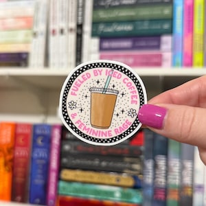 May include: A round sticker with a pink and black border. The sticker features a cartoon illustration of a cup of iced coffee with a straw and the text "Fueled by Iced Coffee & Feminine Rage".