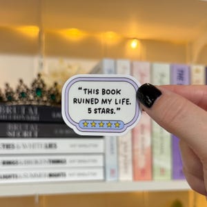 May include: A rectangular sticker with a light purple border and the text "THIS BOOK RUINED MY LIFE. 5 STARS." The sticker has five gold stars at the bottom. The sticker is held in front of a shelf of books.