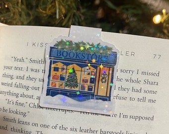 May include: A holographic bookmark featuring a shopfront with a Christmas tree in the window. The shopfront has a sign that reads "BOOKSTORE".