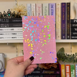 May include: A pink rectangular notepad with iridescent confetti-like accents. The notepad is held in a hand, with a dark nail polish. Bookshelves with books and decorative items are in the background.