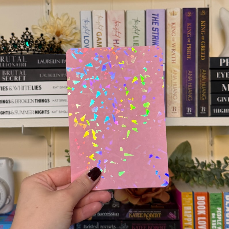 May include: A pink rectangular notepad with iridescent confetti-like accents. The notepad is held in a hand, with a dark nail polish. Bookshelves with books and decorative items are in the background.