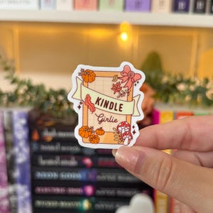 May include: A rectangular sticker with the words "KINDLE Girlie" on a banner. The sticker features orange pumpkins, fall leaves, and a mushroom design. The sticker is held by a person.
