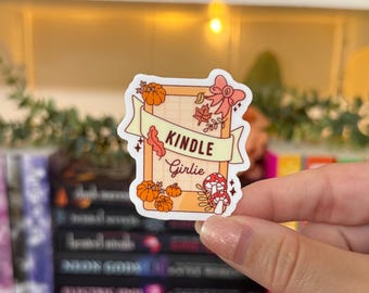May include: A rectangular sticker with the words "KINDLE Girlie" on a banner. The sticker features orange pumpkins, fall leaves, and a mushroom design. The sticker is held by a person.