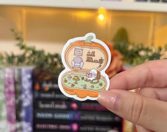 May include: A die-cut sticker featuring a cozy reading scene inside an orange pumpkin. The sticker includes a small character reading in a rocking chair, a bookshelf, and the text "Cozy Reads".