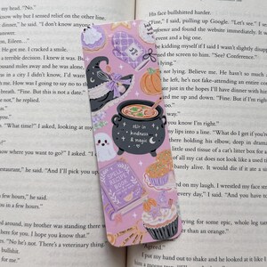 May include: A rectangular bookmark with a light purple background featuring illustrations of a black witch hat, a black cauldron with the text stir in kindness & magic, a spell recipe book, a ghost, a pumpkin, a cupcake, and an oven mitt.