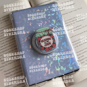 May include: A rectangular e-reader case with a holographic finish featuring a circular phone grip. The grip displays a jar of strawberries with a green lid and the text Reading Is My Jam. The case is overlaid with the text Bookshop Bykendra.