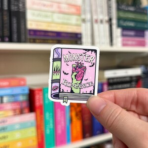 May include: A sticker featuring a pink book with the words "MONSTER ROMANCE." The book has a green spine with black accents and a zombie hand holding a pink heart. Small black bats are scattered around the text.