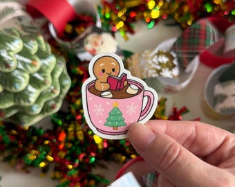 May include: A pink sticker featuring a gingerbread man reading in a mug of hot chocolate. The mug has a green Christmas tree and snowflakes. The background includes Christmas decorations.