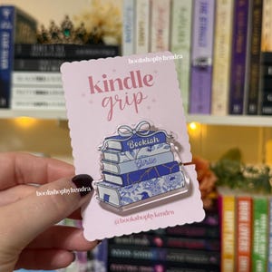 May include: A pink Kindle grip card with the words "kindle grip" in pink script. A clear acrylic book stack pin with the words "Bookish" and "Girlie" is attached. The pin features blue and white floral designs.