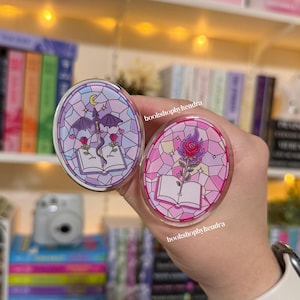 May include: Two oval-shaped acrylic pins with stained glass designs. One features a purple dragon, sword, roses, and an open book. The other has a red rose with flames, an open book, and pink hues. The text "bookshopbykendra" is visible.