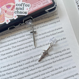 May include: Two silver sword charms with clear crystal beads hanging from a phone case. The phone case is pink and white with the words "coffee and chaos" on it. The phone case is on top of a book with text about a fitness test.