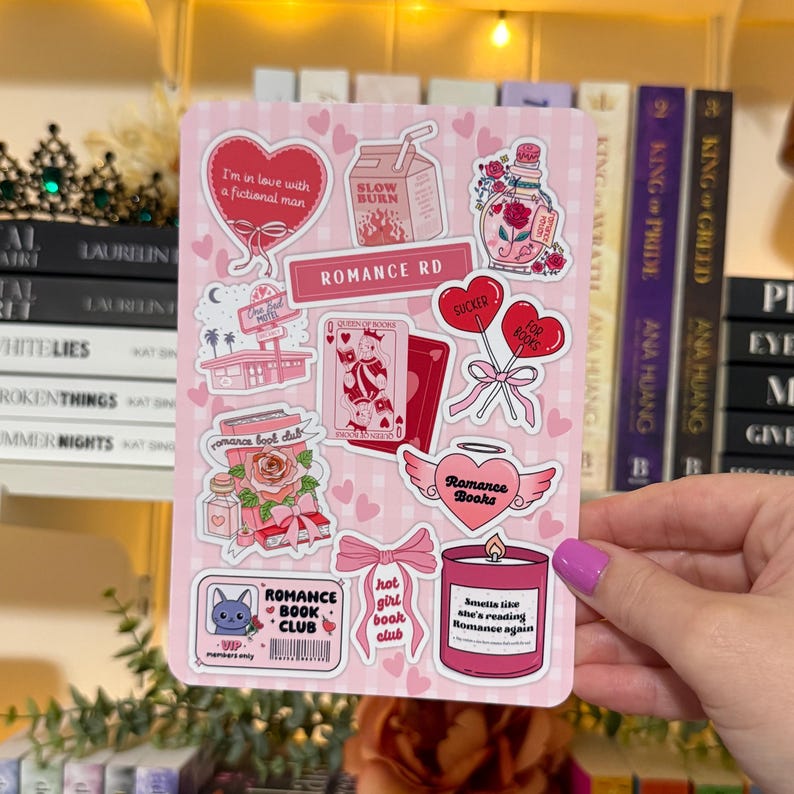 May include: A pink sticker sheet with various romance-themed graphics, including hearts, roses, and the text "Romance Rd." Other text includes "Romance Book Club" and "I'm in love with a fictional man."