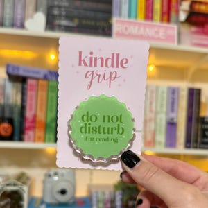 May include: A pink and white Kindle grip with the words "kindle grip" in cursive. The grip has a green, scalloped circle with the words "do not disturb, I'm reading". The background includes a bookshelf with books.