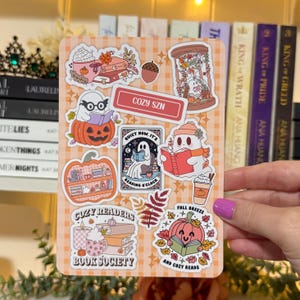 May include: A sticker sheet with fall-themed illustrations. Designs include ghosts reading books, pumpkins, coffee cups, and the text "COZY SZN", "COZY READERS BOOK SOCIETY", "FALL BREEZE AND COZY READS", and "QUIET NOW IT'S READING O'CLOCK". The background is a light orange plaid.