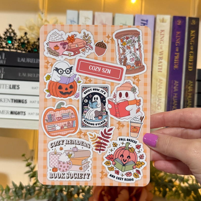 May include: A sticker sheet with fall-themed illustrations. Designs include ghosts reading books, pumpkins, coffee cups, and the text "COZY SZN", "COZY READERS BOOK SOCIETY", "FALL BREEZE AND COZY READS", and "QUIET NOW IT'S READING O'CLOCK". The background is a light orange plaid.
