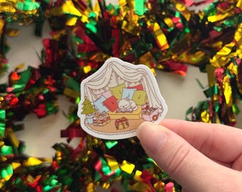 May include: A sticker featuring a cozy scene with a white cat sleeping on a bed of pillows inside a tent. Presents and a Christmas tree are nearby. The sticker is held in a hand, with colorful tinsel in the background.