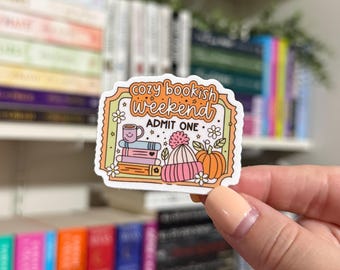 May include: A sticker with a whimsical illustration of a cozy bookish weekend. The sticker features a stack of books, a mug of hot cocoa, a cozy hat, and a pumpkin. The text on the sticker reads "Cozy Bookish Weekend" and "Admit One".