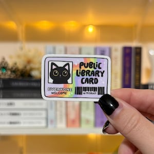 May include: A rectangular sticker featuring a black cat illustration on a lavender background. The sticker reads "Public Library Card" with a rainbow gradient and the words "Everyone Welcome". A barcode and the number 67938001 are also present.