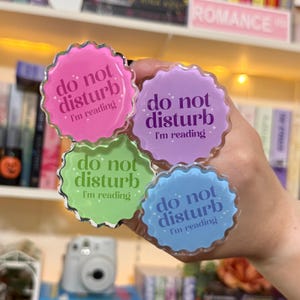 May include: Four colorful, scalloped-edge acrylic pins with the text "do not disturb, I'm reading" in white script. The pins are in pink, purple, green, and blue. The background includes a bookshelf and the word "ROMANCE".