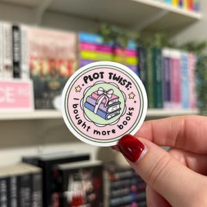 May include: A round sticker with a pink and green background. The sticker says "Plot Twist: I bought more books" with a stack of books and a pink bow on top.
