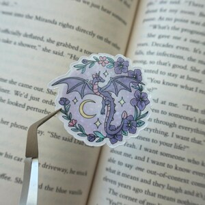 May include: A circular sticker featuring a purple dragon with wings spread, centered above a yellow crescent moon. The dragon is surrounded by a wreath of purple and pink flowers with green leaves against a light purple background with small stars.
