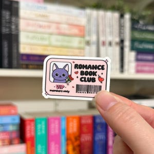 May include: A pink and white sticker with a cartoon cat holding flowers. The sticker says "Romance Book Club" and "VIP members only".