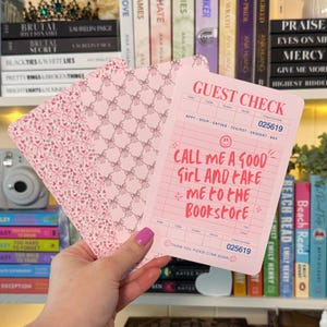 May include: Pink guest check with the text "CALL ME A GOOD GIRL AND FAKE ME TO THE BOOKSTORE." The check has a bow pattern on the back and a check number of 025619. The background includes books and a camera.