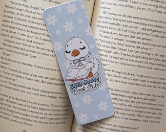 May include: A light blue bookmark with a cartoon goose reading a book, with the text "Silly Goose Book Club." The bookmark has a floral pattern and is set against the backdrop of an open book.