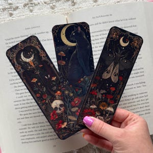 K&ouml;nnte beinhalten: Drei dekorative Lesezeichen mit dunklen, gotisch inspirierten Illustrationen. Eines zeigt eine schwarze Katze unter einem Halbmond, ein anderes eine Motte und das dritte einen Totenkopf. Florale und Pilzdetails schm&uuml;cken die Designs.