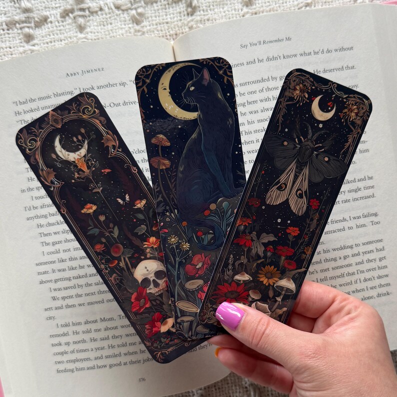K&ouml;nnte beinhalten: Drei dekorative Lesezeichen mit dunklen, gotisch inspirierten Illustrationen. Eines zeigt eine schwarze Katze unter einem Halbmond, ein anderes eine Motte und das dritte einen Totenkopf. Florale und Pilzdetails schm&uuml;cken die Designs.
