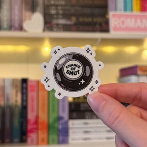 Könnte beinhalten: Ein weißer Aufkleber mit einem Design einer schwarzen und grauen Kristallkugel. Der Text "CHANCE OF SMUT" befindet sich in der Kristallkugel. Der Aufkleber hat eine kreisförmige Form mit vier sternförmigen und vier kleinen quadratischen Designs am Rand.