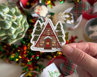 May include: A sticker featuring a brown and red A-frame cabin with green trees and a snowy base. The cabin has a red door and windows, with the text "Book Nook" above the door. The sticker is surrounded by Christmas decorations.