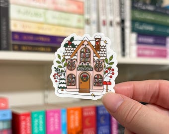 May include: A pink and white illustrated sticker of a house with a sign that reads "Holly Jolly Bookstore". The house has a red door, a wreath, and windows with books inside. There are green trees and red berries around the house.